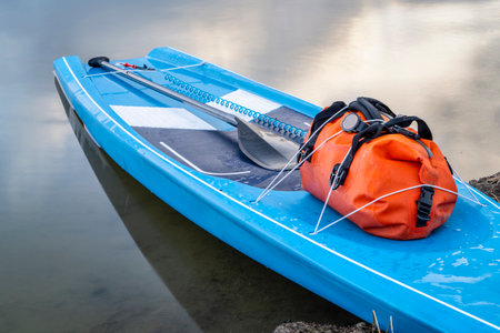 touring stand up paddleboard with a waterproof duffel, paddle and safety leash on a lake shore,...