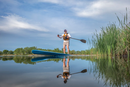 senior male paddler is paddling  a stand up paddleboard on a calm lake in spring, frog perspective...