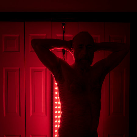 silhouette of a senior athletic, man torso standing in front of the red light therapy panel, home setup for treatmentの写真素材