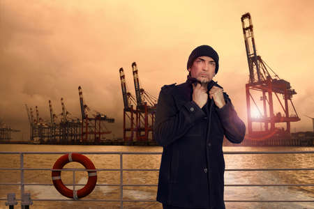 A sailor stands on the deck of a ship with his collar turned up, the cranes in the harbor can be...