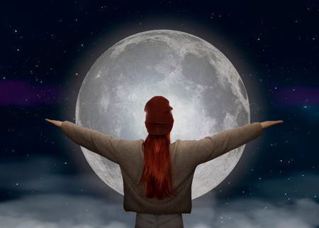 A young woman with red hair and a knitted hat looks to the moon with her arms raisedの写真素材