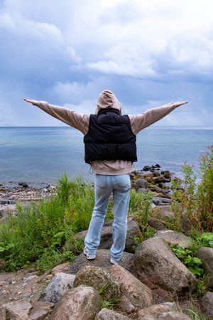 A woman stands on a rock by the sea under a cloudy sky and raises her armsの写真素材