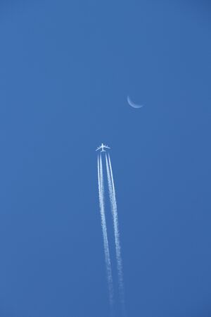 Silhouette of passenger plane with condensation trails against cloudless blue sky with waning moon