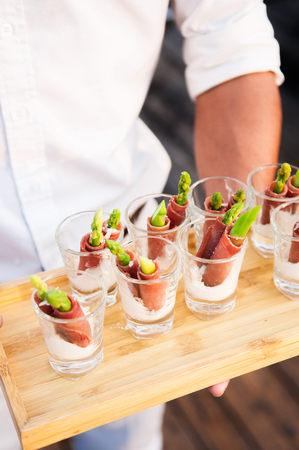 Canape from asparagus and dried ham with creamy sauce in glass serving by waiter on wooden trayの写真素材