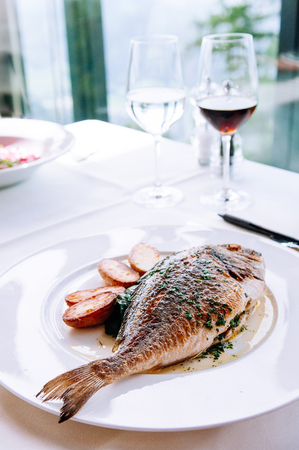 Grilled sea bream fish with butter sauce, parsley and baked potatoes on white plate. Close up shotの写真素材