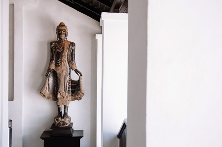 Beautiful old carved wood standing Buddha sculpture, Lanna northern Thai style antique Buddha with white wall backgroundの写真素材