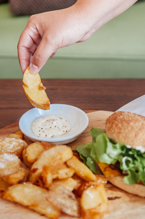 Beef cheeseburger and hand picking fried potato chunks with ranch dipping sauce on wood plate topside view shotの写真素材