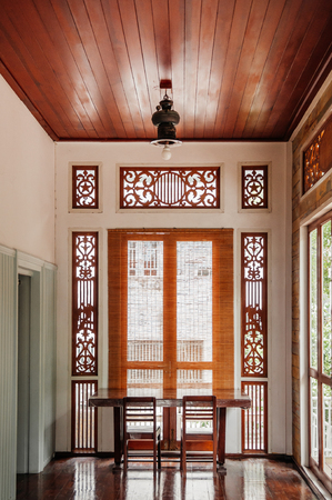 AUG 4, 2014 Bangkok, Thailand - Oriental Thai-Chinese Oriental vintage wooden desk and chair at high ceiling hallway in old house with carved wood windows and brown wood floor with natural light from windowのeditorial素材