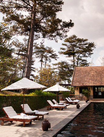 FEB 25, 2014 Dalat, Vietnam - Tropical Colonial resort style swimming pool in pine forest under evening sunlight in late winter with reflection on water surface and white pool beds and umbrellasのeditorial素材