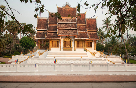 APR 4, 2019 Luang Prabang, Laos - Luang Prabang Royal Palace Museum and Hor Prabang temple hall seen throuhg tree branches with empty street in morningのeditorial素材