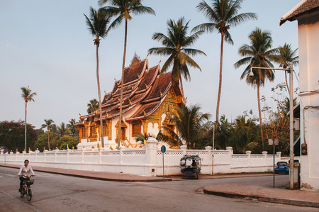 APR 4, 2019 Luang Prabang, Laos - Luang Prabang Royal Palace Museum and Hor Prabang temple hall under coconut tree with Tuk Tuk park at wall side and morning quiet streetのeditorial素材