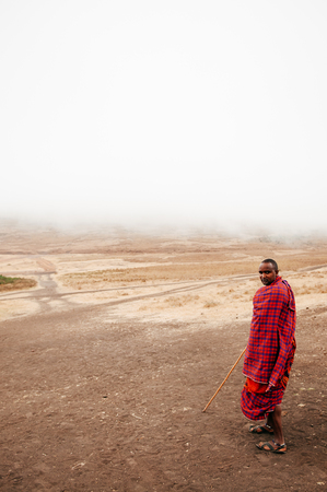 JUN 24, 2011 Serengeti, Tanzania - African Masai or Maasai tribe man in red cloth standing alone in empty dusty land on cold foggy day. Ngorongoro Consevation, Serengeti Savanna forest in Tanzania.のeditorial素材