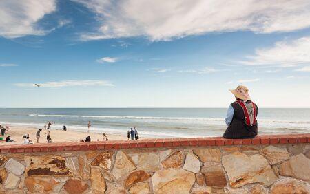 Rear view of lady sitting on a wall in front of the beach in Adelaide and observing life on the beach of Australiaの写真素材
