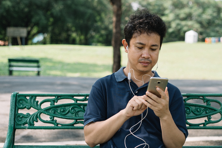 Asian man with smartphone sitting on the bench in the parkの写真素材