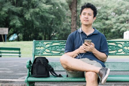 Asian man with smartphone sitting on the bench in the parkの写真素材