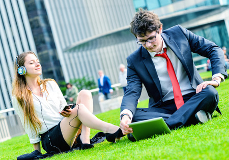 Successful Junior executives dynamics laying down on green grass near a modern glass office building, listening music and using laptopの写真素材
