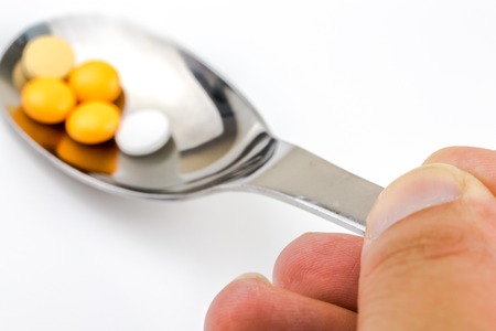 A hand holding a silver spoon with yellow and white medical pills against white background