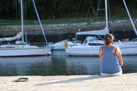 A young woman sitting at the harbor and talking on her phone looking at the ships in the waterの写真素材