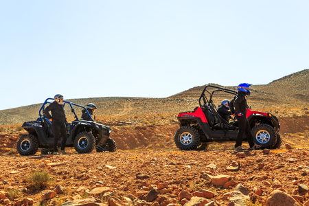 Merzouga, Morocco - February 21, 2016: Four riders from two quad ATVs stand beside their vehicles on rocky terrain and survey the path ahead. Overhead is an overcast sky and in the distance a range of large hills can be seenのeditorial素材