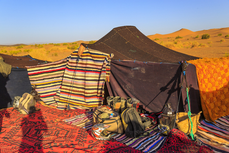 Merzouga, Morocco - February 25, 2016: The shadow of a photographer falls over the colorful rugs and blankets surrounding a black nomads tent in the Sahara desert under a bright blue sky.のeditorial素材