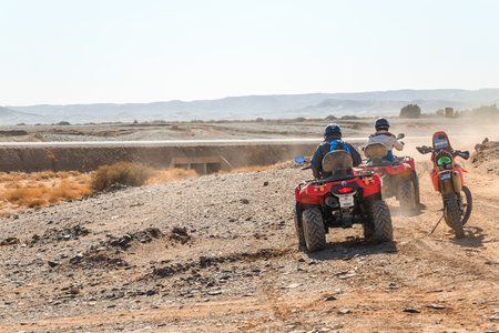 Merzouga, Morocco - February 25, 2016: Two riders on quad ATVs wait beside a riderless motorbike on rocky terrain and watch an empty highway which crosses their path. Overhead is an overcast sky and in the distance a range of mountains can be seen.のeditorial素材