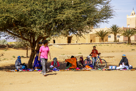 Tinzouline, Morocco - February 26, 2016: Berber vendors sit in the shade of a spreading tree in a moroccan village. Their goods are spread out on the ground in front of them in the traditional manner of a marketplace, or souq, as a tourist walks byのeditorial素材