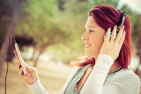 Portrait of an attractive smiling redhead woman listening to music on her phone with a headset with...