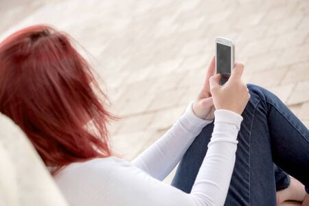 Pretty smiling redhead woman sending messages on her cell phone in a park. . Focus on her phone
