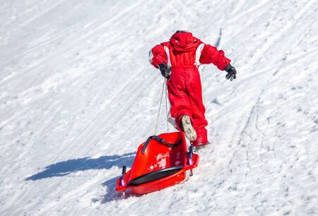La Mongie, France - March 21, 2019:Back view of child in red outwear pulling sledge and running up snowy slope while spending time on ski resortの写真素材