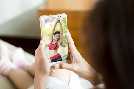 Young woman sit on sofa watching demonstration of Yoka on her smartphone.の写真素材