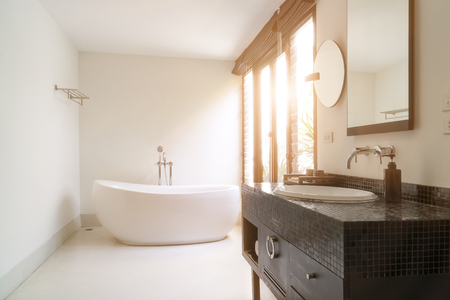 Modern bathroom interior with white oval bathtub with bright sunlight through window with a view of green trees.の写真素材
