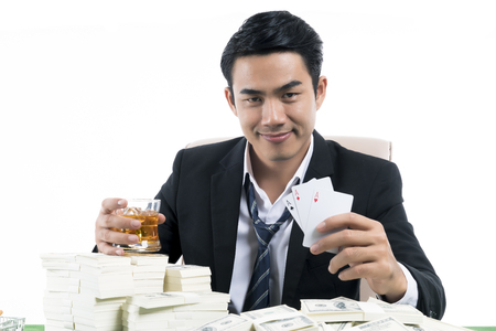 Portrait the young rich man holding whiskey glass and show winning with cards and stack of dollars on green table, white background with business casino conceptの写真素材