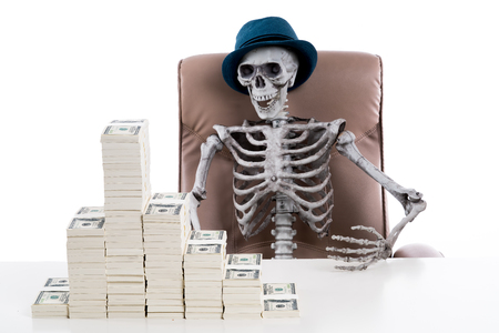 A Human skeleton wearing blue hat and sitting in a big chair with stack of dollars on white table, rich man concept
の写真素材