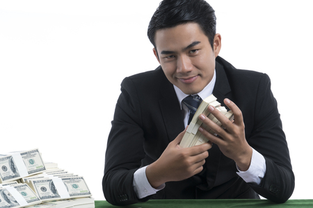 A Handsome man is smiling and holding piles of banknotes in hands, has a lot of dollars placed nearby, on green table, white background and copy space, casino business concept
の写真素材