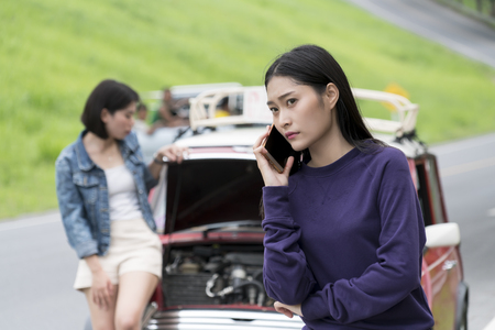 The young female calling autoservice with broken down car during travel and her friend standing near red car at side streetの写真素材