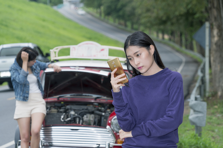 The young female see at her mobile phone in hand and using calling mechanic service the broken down car during travel with her friend standing near red car at side streetの写真素材