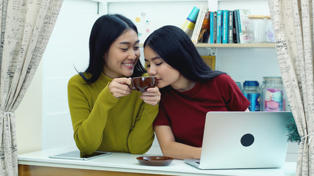 Two asian lovely teenagers using laptop computer to working, they are joyful to talking and Smell of coffee during typing on keyboard at the living room in house, Teenager lifestyle conceptの写真素材