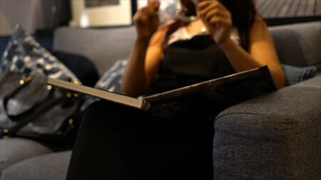 Middle-aged woman sitting on sofa get to wear glasses and flib to read a book at nightの写真素材