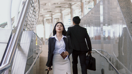Asian young woman walks along a baggage with haste reading the signpost to find destination. Blurred back of businessman walk going in the opposite directionの写真素材