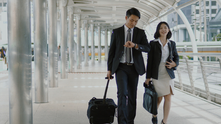Selective focus at Asian businessman dragging luggage and checking time with a wristwatch, pretty businesswoman walking and talking together in rush hourの写真素材