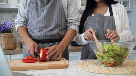 Young husband help pregnant use knife to slicing red sweet pepper,  and put into glass of vegetables salad and mix themの写真素材