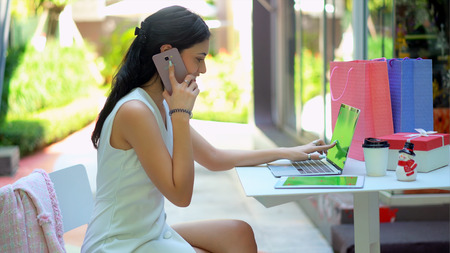 The young pretty woman looks very happy after talking on smartphone and point to green screen of laptop, has tablet with green screen and shopping bag nearlyの写真素材