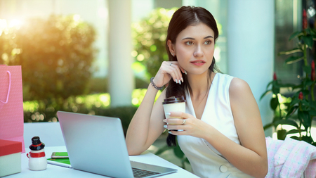Close up with the pretty woman looking fresh after drink beverage in her hand, has laptop and shopping bag on tableの写真素材