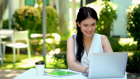 The pretty woman sitting and working on laptop with happiness in the garden, On her desk has equipment technology with smartphone, tablet and laptop computer with green screen to workの写真素材