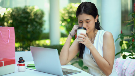 Close up the pretty woman looking fresh after smell of beverage in her hand, has laptop and shopping bag on tableの写真素材