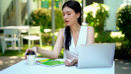 Close up the young woman pick up a coffee cup on table in garden, has laptop, smartphone , tablet  and shopping bag placed nearlyの写真素材