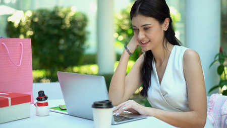 Close up the young pretty woman sitting and rest the chin on her hand, looking to laptop and smile with cheerful, has tablet with green screen and shopping bag nearlyの写真素材