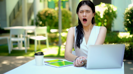 The young woman open her mouth to yawn drowsy while she works at outside the office with laptop on the table in gardenの写真素材