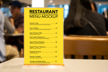 Mock up vertical menu with clipping path on table, yellow screen empty space in acrylic for list of menu at restaurant, copy space in right and blurred people sitting in backgroundの写真素材