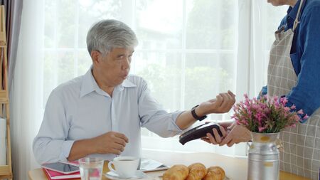 Asian senior man sitting and using tablet in coffee shop, when waiter male come in the old man...